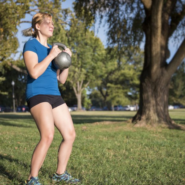 Young fit woman doing squats with heavy kettlebell in green outdoor park
